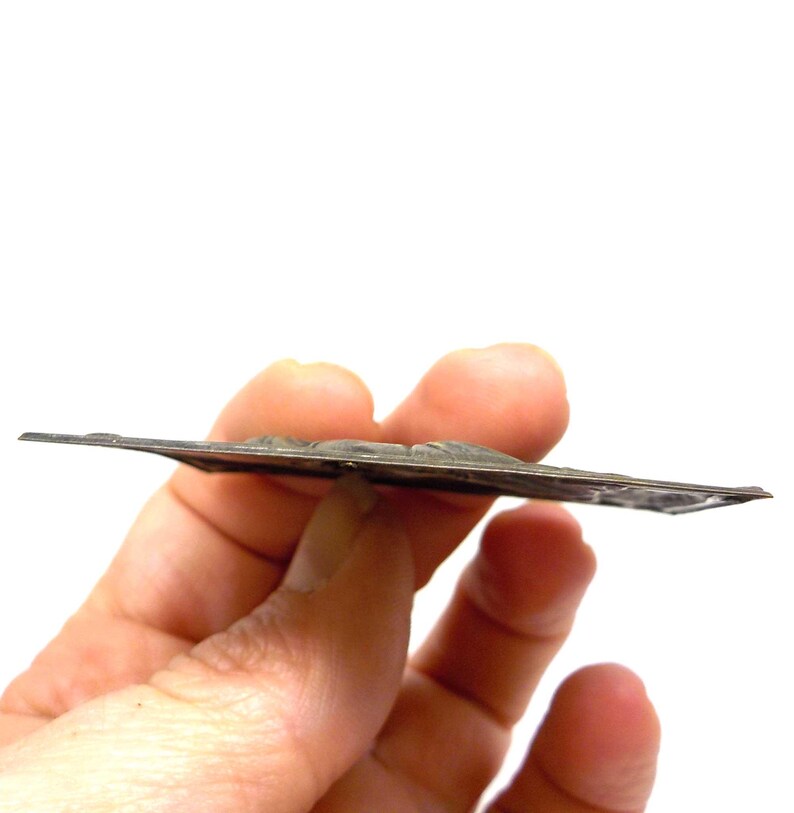 May include: A silver-toned, rectangular metal piece with a raised relief design. The object is held in a hand against a white background. The metal has a slightly tarnished appearance, suggesting age or wear.
