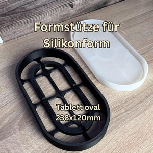 Könnte beinhalten: Eine schwarze ovale Formstütze für Silikonformen und eine weiße ovale Silikonform. Der Text auf dem Bild lautet Formstütze für Silikonform und Tablett oval 238x120mm. Der schwarze Rahmen hat eine gitterartige Struktur, um die Silikonform zu halten.