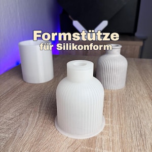 Könnte beinhalten: Drei weiße Formen werden auf einer Holzoberfläche gezeigt. Die Formen umfassen einen hohen, glatten Zylinder, eine geriffelte Vase und eine geriffelte, glockenförmige Form. Der Text oben lautet "Formstütze für Silikonform."