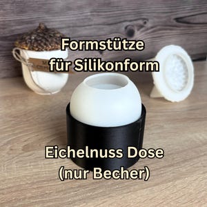 Könnte beinhalten: Eine schwarze zylindrische Formstütze mit einer weißen, kuppelförmigen Silikonform. Der Text Formstütze für Silikonform und Eichelnuss Dose (nur Becher) ist eingeblendet. Im Hintergrund ist ein weißer Behälter in Eichelform zu sehen.