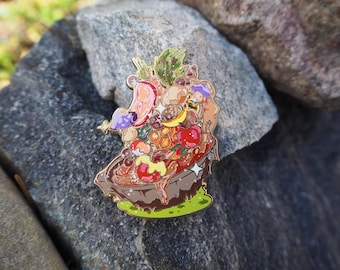 May include: A colorful enamel pin featuring a whimsical design of a dessert or food arrangement. The pin includes various elements like fruits, honeycombs, and other food items, with a gold outline and a green base. The pin is set against a natural stone background.