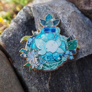 Pode incluir: Um pin detalhado em esmalte com um design de frasco de poção azul com elementos florais e aquáticos. O pin tem um contorno dourado e está sobre um fundo de pedra natural. O design inclui uma flor branca central e tons de azul.