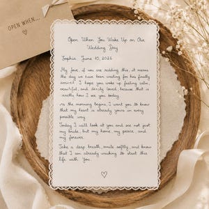 Pode incluir: Uma carta de casamento manuscrita em papel texturizado com uma delicada borda de renda, colocada em uma bandeja de madeira. A carta inclui o texto "Open When You Wake Up on Our Wedding Day" e uma mensagem pessoal. Um envelope com o texto "OPEN WHEN..." também é visível.