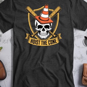 May include: Dark gray t-shirt featuring a skull wearing an orange and white traffic cone, crossed baseball bats, and the text "HOIST THE CONE" in a banner. The design is set against a yellow shield.