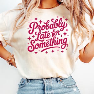 May include: Cream-colored t-shirt with the phrase "Probably Late for Something" in a cursive font, surrounded by starburst graphics. The text and stars are in a deep pink color. The shirt has rolled-up sleeves and is paired with blue jeans.
