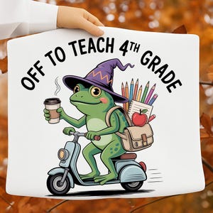 May include: A white fabric surface featuring a graphic of a green frog wearing a purple witch hat, riding a light blue scooter. The frog holds a coffee cup and carries a backpack with pencils and an apple. Text reads: OFF TO TEACH 4TH GRADE.