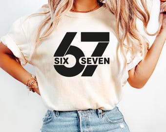 May include: Cream-colored t-shirt with the number 67 in bold black font. The word "SIX" is centered within the zero of the 6, and "SEVEN" is below the 7. The shirt is worn with blue jeans.