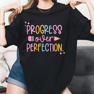 May include: Black t-shirt with the phrase "Progress Over Perfection" in colorful, playful lettering. The design includes a pink pencil, a yellow pencil, and star accents. A casual, comfortable tee.