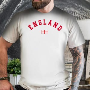 Könnte beinhalten: Ein weißes T-Shirt mit kurzen Ärmeln, das auf der Brust das Wort ENGLAND in einer fetten, bogenförmigen roten Schrift zeigt, platziert direkt über einer kleinen Grafik der rot-weißen englischen Flagge in der Mitte des Kleidungsstücks.