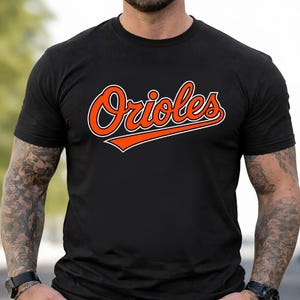 May include: Black t-shirt featuring the word "Orioles" in orange script with a white outline and an orange underline. The shirt is a crew neck style.