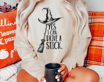 May include: Beige sweatshirt with a black witch hat and broom graphic. The text reads "YES I CAN DRIVE A STICK." The design is a Halloween or fall-themed graphic. The person is holding a black mug.