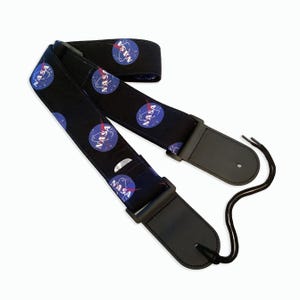 May include: Black guitar strap with a repeating pattern of the NASA logo in blue, red, and white on a black background. The strap has black leather ends with adjustable straps.