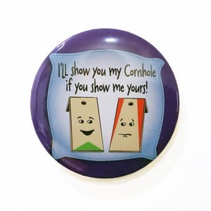 May include: Purple round button with a white background and the text "I'll show you my Cornhole if you show me yours!" with two cartoon cornhole boards with faces.