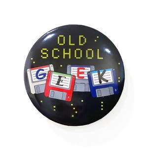 May include: A black button with the text "OLD SCHOOL GEEK" in yellow pixelated letters. The letters are surrounded by four colorful floppy disks with the letters "G", "E", "E", and "K" on them.