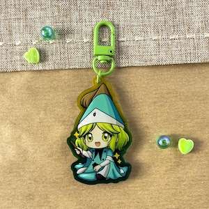 Può includere: Un portachiavi verde e verde acqua in stile anime con un personaggio in un abito da strega. Il portachiavi ha un fermaglio verde ed è accompagnato da piccole perle verdi e un ciondolo a forma di cuore. Lo sfondo è di colore beige neutro.
