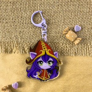 Può includere: Un portachiavi in acrilico trasparente con un personaggio dei cartoni animati con capelli viola, un abito rosso e oro e un cappello marrone. Il portachiavi ha una chiusura viola. Sono presenti anche due caramelle e due decorazioni a forma di cuore viola.