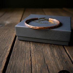 Puede incluir: Una pulsera de puño de cobre martillado descansa sobre una caja de regalo gris, sobre un fondo de madera rústica. La pulsera tiene una superficie texturizada y un cálido tono marrón rojizo. La caja es gris oscuro y la madera tiene un aspecto envejecido.