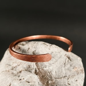 Op de afbeelding: Een koperen manchetarmband met een getextureerd oppervlak. De open armband wordt getoond op een lichtgrijze steen tegen een donkere achtergrond. Dit minimalistische sieraad is een stijlvol accessoire.