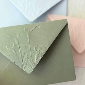 May include: A sage green envelope featuring a raised embossed design of three calla lily flowers and leaves on the triangular flap. The envelope is made of heavy cardstock and is positioned in front of a light blue envelope and a pale pink envelope.