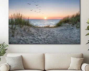 May include: A large canvas print depicting a serene beach scene at sunset. The image features a sandy path leading to the ocean, with tall grass on either side. The sky transitions from blue to orange, with birds flying above the water. The print is displayed above a beige sofa.