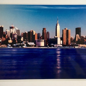 May include: A vintage photograph of the New York City skyline with the Empire State Building in the centre. The photo is taken from across the water, with the city's skyscrapers reflected in the water.
