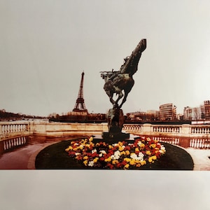 May include: A bronze statue of a man on horseback stands in a park with the Eiffel Tower in the background. The statue is surrounded by a bed of colourful flowers.