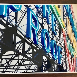 May include: A close-up view of a large, blue neon sign that reads "The Roof", mounted on a metal framework. The sign is illuminated and visible against a bright, white sky.