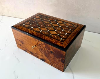 May include: A rectangular wooden box with a decorative inlaid top featuring a geometric pattern of diamonds and squares. The box has a burl wood grain and a gold-colored keyhole.