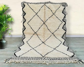 May include: A white and black Moroccan rug with a diamond pattern. The rug has a fringe border and is made of wool.