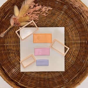 May include: Four pastel-coloured rectangular hair clips in a wicker basket. The clips are in shades of pink, purple, orange, and white.