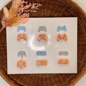 May include: A set of 10 different coloured resin game controller pins. The pins are in the shape of various game controllers, including the Nintendo 64, Super Nintendo, and Playstation. The pins are arranged in three rows on a white background.