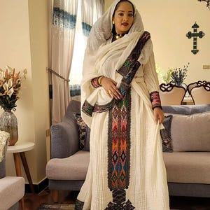 Puede incluir: Una mujer con un vestido blanco de manga larga y un velo a juego. El vestido presenta patrones bordados coloridos en el centro y en las mangas. La mujer lleva joyas doradas.