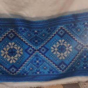 Puede incluir: Textil con un detallado patrón bordado en azul y blanco. El diseño presenta formas geométricas y motivos florales, creando un llamativo contraste visual con la tela blanca. El bordado probablemente está cosido a mano, mostrando artesanía.