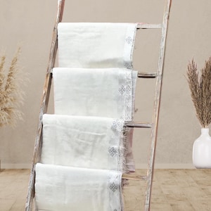 May include: Four folded white blankets with a decorative border are displayed on a rustic wooden ladder. The blankets are neatly arranged, showcasing their texture and design. The scene is set against a neutral background, with decorative plants in the foreground.