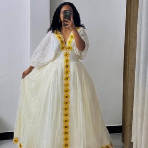 May include: A white, floor-length dress with a V-neckline and long, puffy sleeves. The dress is adorned with yellow floral embroidery along the neckline, down the front, and around the hem. The fabric appears to be lightweight and flowing.