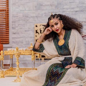 Puede incluir: Una mujer con un vestido tradicional color crema con bordados verdes y multicolores. Lleva joyas de oro, incluyendo un collar, pendientes y un adorno para la cabeza. Una mesa dorada está en primer plano.