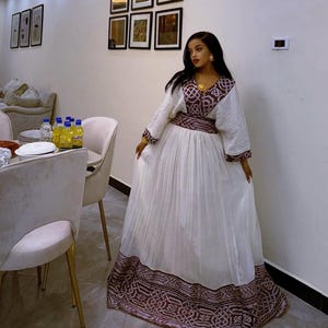 Puede incluir: Un vestido blanco largo hasta el suelo con mangas largas y una falda plisada. El vestido presenta intrincados bordados marrones y morados alrededor del escote, la cintura y el dobladillo. La mujer lleva joyas de oro.