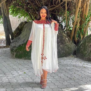 Puede incluir: Un vestido blanco con detalles bordados en rojo. El vestido tiene mangas largas y transparentes y una silueta fluida. El escote y los puños presentan intrincados patrones rojos. El vestido es hasta la rodilla y se lleva con sandalias de tiras.