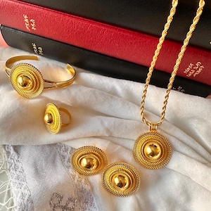Könnte beinhalten: Ein goldfarbenes Schmuckset mit Armband, Ring, Ohrringen und einer Halskette mit Anhänger. Jedes Stück hat eine zentrale Kuppel, die von einem strukturierten, kreisförmigen Design umgeben ist. Das Schmuckstück liegt auf weißem Stoff.