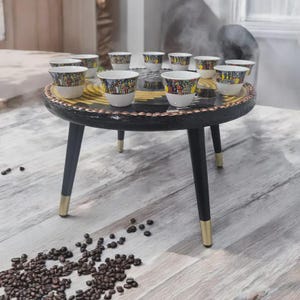 Puede incluir: Una pequeña mesa redonda negra con patas doradas sostiene una colección de pequeñas tazas de café blancas. Cada taza presenta una ilustración colorida y detallada en su exterior. Los granos de café están esparcidos en el suelo de madera.