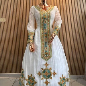 May include: A white dress with intricate embroidery in teal, gold, and orange. The dress features long sleeves and a full skirt, with detailed patterns along the neckline, bodice, and hem. The dress is displayed on a mannequin.