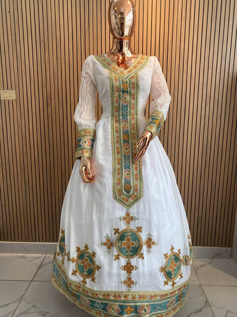 May include: A white dress with intricate embroidery in teal, gold, and orange. The dress features long sleeves and a full skirt, with detailed patterns along the neckline, bodice, and hem. The dress is displayed on a mannequin.