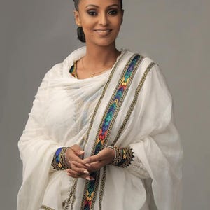 Puede incluir: Una mujer con una prenda blanca con adornos bordados coloridos. El adorno presenta un patrón geométrico con detalles azules, verdes, amarillos y rojos. La mujer tiene el pelo oscuro recogido en un moño y lleva un collar de oro.