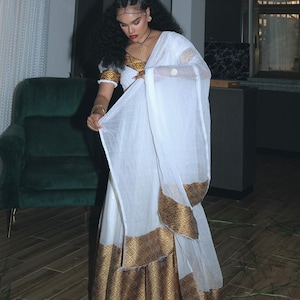 Puede incluir: Un vestido blanco con adornos y detalles dorados. El vestido presenta una falda larga y un chal a juego. El adorno dorado tiene un patrón geométrico. Las mangas son cortas y el escote está adornado con oro.