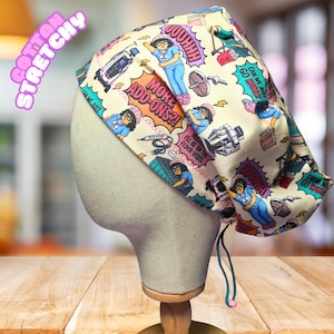 May include: A scrub cap featuring a cream-colored fabric with a repeating cartoon print of a character in blue clothing, medical equipment, and speech bubbles with text including "MORE ADD-ONS?!" and "OUHHH". The cap has a teal drawstring.