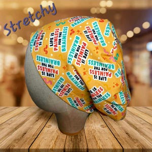 May include: A yellow scrub cap with the text "Life is Painless for the Brainless" in white, blue, and red. The cap has a repeating pattern of the phrase and various shapes. The word "Stretchy" is written in blue.
