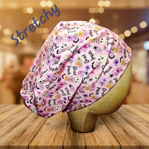 May include: A pink, stretchy surgical scrub cap with a repeating pattern of skulls, spiders, flowers, and the words "Dead Inside." The cap is displayed on a wooden mannequin head.