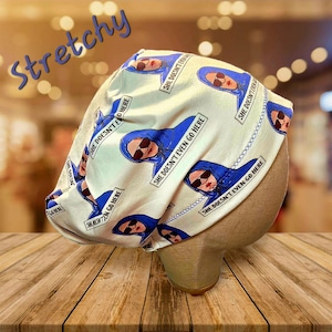 May include: A white and blue patterned scrub cap with the text "SHE DOESN'T EVEN GO HERE". The cap features a repeating design of a woman in a blue hoodie and sunglasses. The word "Stretchy" is written in blue script.