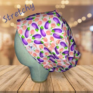 May include: A pink and white scrub hat with a pattern of purple eggplants and peach slices. The hat is made of a stretchy fabric and has a small label on the side.