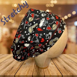 May include: Black and red patterned scrub cap with a cartoon bat design, white stars, and red accents. The word "Stretchy" is written in blue script. The cap is displayed on a mannequin head.